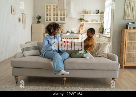Felice figlio africano americano bambino sorprendente mamma il giorno delle madri a casa, dando dono alla mamma Foto Stock