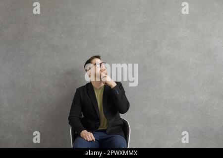Serio giovane uomo in occhiali seduti in sedia, guardando verso l'alto e pensando a qualcosa Foto Stock