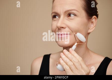 Donna di mezza età che ama massaggiare il viso con rullo al quarzo rosa Foto Stock