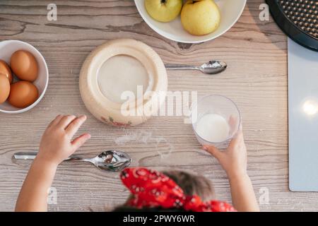 Mani di bambina in fascia rossa cuocere torta di mele in cucina. Il bambino mette lo zucchero nel misurino. I bambini aiutano sui lavori domestici. Cibo da cucina per bambini Foto Stock