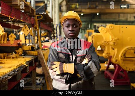 Giovane operaio afro-americano serio in elmetto protettivo e abbigliamento da lavoro guardando la macchina fotografica mentre si trova in piedi in un impianto industriale Foto Stock