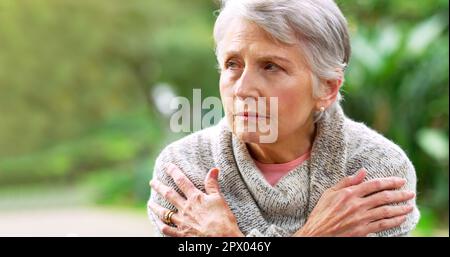 È un po' freddo oggi. una donna anziana spensierata seduta su una panchina mentre contempla fuori in un parco Foto Stock