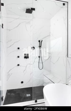 Una bella cabina doccia con pareti piastrellate in marmo, pavimento in piastrelle circolari nere e rubinetti neri. Foto Stock
