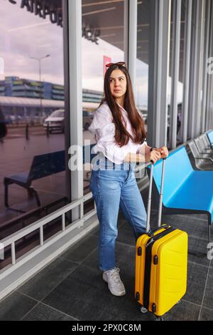 Giovane ragazza traveller vestita casualmente con valigia gialla in attesa nella sala del terminal dell'aeroporto. Focus sulla donna. Foto Stock