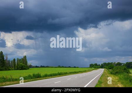 Autostrada vuota, strada asfaltata attraverso i campi. Cielo drammatico, pioggia all'orizzonte in lontananza. La tempesta sta arrivando. Concetto di paesaggio. Foto Stock