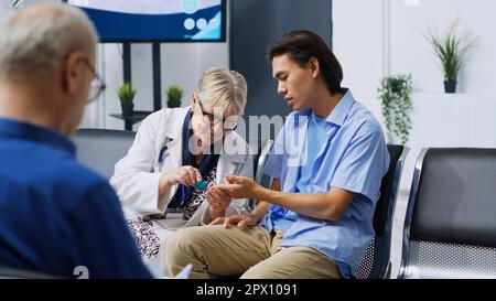 Medico che prende paziente test del livello di insulina usando glucometro medico nella zona di attesa dell'ospedale. Medico anziano che esegue un esame del diabete per misurare il glucosio dal campione di sangue. Servizio di medicina Foto Stock