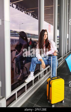 Giovane ragazza traveller vestita casualmente con valigia gialla in attesa nella sala del terminal dell'aeroporto. Focus sulla donna. Foto Stock