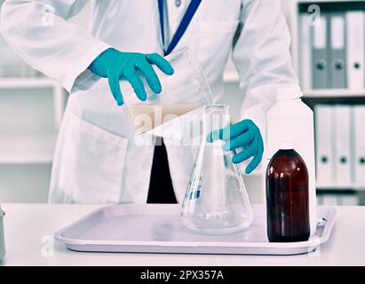 Preparazione del suo esperimento...una scienziata femminile irriconoscibile che trasferisce un liquido da un bicchiere a una beuta conica mentre lavora in laboratorio Foto Stock