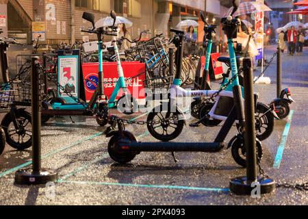 Osaka, Giappone - 29 aprile 2023: Forte pioggia su mini bici elettriche condivise e scooter parcheggiati sulla strada cittadina Foto Stock