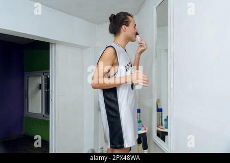 Latina giovane di sesso non binario in piedi nella sua stanza di fronte allo specchio, preparandosi, finendo di applicare il trucco. Foto Stock