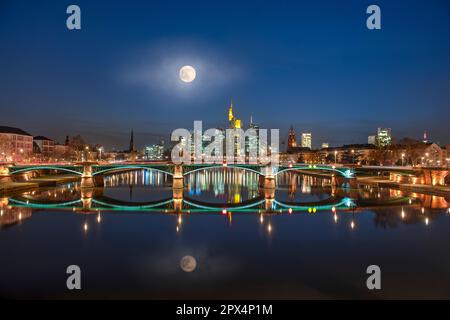 La luna piena si riflette nel fiume meno con lo skyline serale di Francoforte sul meno illuminato artificialmente Foto Stock