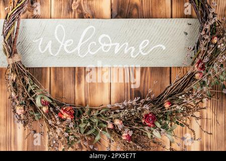 Fiore asciutto, che si vagliano strettamente tessuto corona con stringa e nome bordo Benvenuto. Rustica porta di legno marrone e spianatura sullo sfondo. Celebrazione, preparazione Foto Stock