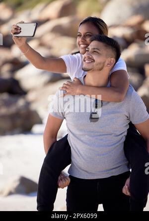 Spiaggia, selfie e piggy back coppia, sorridere con smartphone in vacanza romantica estate al mare. Romanticismo, uomo e donna con telefono, amore e vacatio Foto Stock