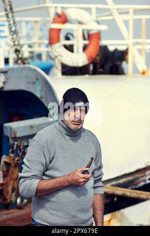 Barca del pescatore, ritratto dell'uomo maturo e tubo fumante vicino all'acqua di mare ad un porto per la pesca. Anziano, oceanico e lavoratore portuale senior seduto in barca Foto Stock