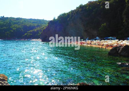 Budva, Montenegro, spiaggia di Mogren. Agosto 15 2022 Spiaggia di sabbia e ciottoli vicino a alte scogliere. Ombrelloni, caffè, sedie a sdraio, sdraio. Le persone si rilassano Foto Stock