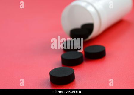 Diverse compresse nere cadono dal vaso di plastica sulla superficie rossa. Immagine di sfondo su argomenti medici e farmaceutici. Carbone attivo Foto Stock