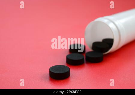 Diverse compresse nere cadono dal vaso di plastica sulla superficie rossa. Immagine di sfondo su argomenti medici e farmaceutici. Carbone attivo Foto Stock