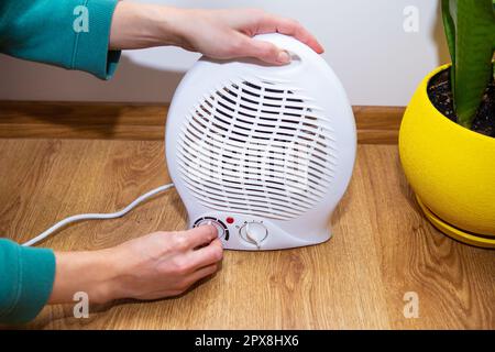 La ragazza tiene le mani vicino al riscaldatore del ventilatore di plastica e le riscalda le mani, regolando la temperatura nel paese Foto Stock