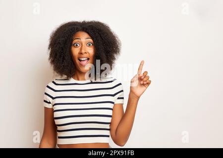 Una foto al coperto di una donna felice dai capelli ricci punta il dito da parte per copiare lo spazio, vestito con una t-shirt a righe, si posa su una parete bianca. Foto di alta qualità Foto Stock