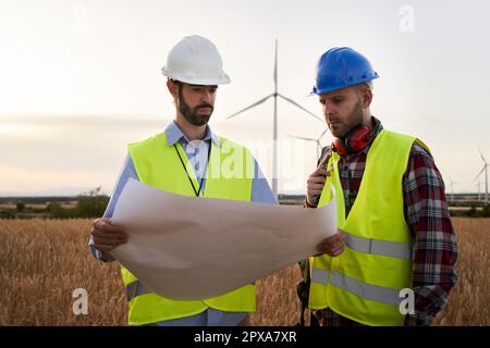Due ingegneri del vento che indossano giubbotti e caschi guardano i piani del sistema di turbine eoliche sul campo. Foto Stock
