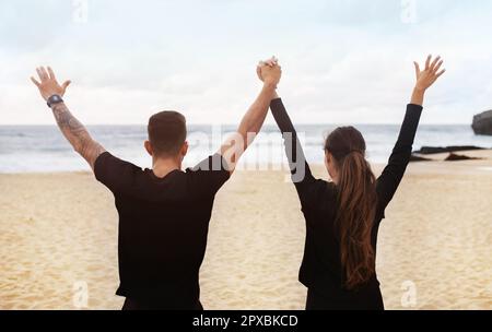 Coppia sportiva felice godendo la pausa di workout, tenendo e alzando le mani, guardando l'oceano, vista posteriore Foto Stock