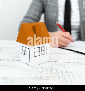Donna d'affari seduta e scritta nel notebook. Paper house sulla scrivania. Foto Stock