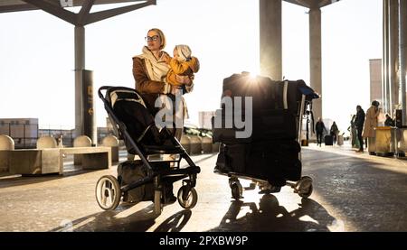Motherat viaggia con il bambino, camminando, spingendo passeggino e carretto bagagli davanti alla stazione del terminal aeroportuale Foto Stock