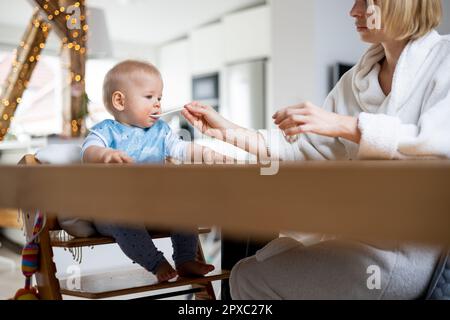 La madre indossa un comodo cucchiaio da bagno che allatta il bambino in poltrona con purea di frutta al tavolo da pranzo a casa. Introduti di alimenti solidi per bambini Foto Stock