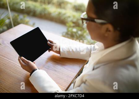 Giovane donna nera in tuta bianca e bicchieri siede al tavolo di legno, guarda il video su un tablet a schermo vuoto Foto Stock