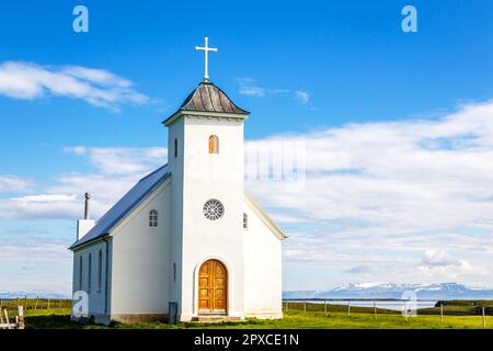 Flateyjarkirkja bianca chiesa luterana con prato in primo piano e fiordo di mare con cielo blu e montagne sullo sfondo, Flatey Island, Islanda Foto Stock