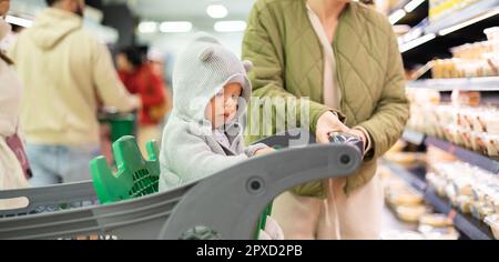 Casualy vestito madre scelta prodotti nel reparto del supermercato negozio di alimentari con il suo bambino bambino bambino bambino nel carrello della spesa Foto Stock