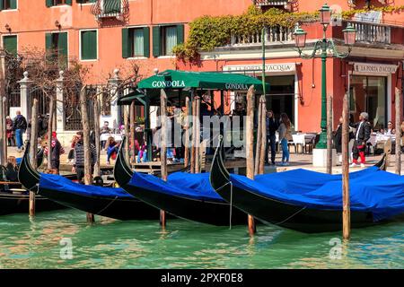 Gondole legate alle colonne di legno sul Canal Grande come persone che camminano sulla strada sullo sfondo a Venezia, Italia. Foto Stock