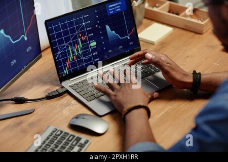 Mani di un giovane intermediario afroamericano che digita sulla tastiera del notebook e analizza il grafico finanziario sullo schermo mentre sei seduto sul posto di lavoro Foto Stock