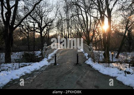 Una pittoresca scena invernale caratterizzata da un Central Park pieno di neve, con un ponte stagionato e alberi che si stagliano contro uno splendido cielo al tramonto Foto Stock