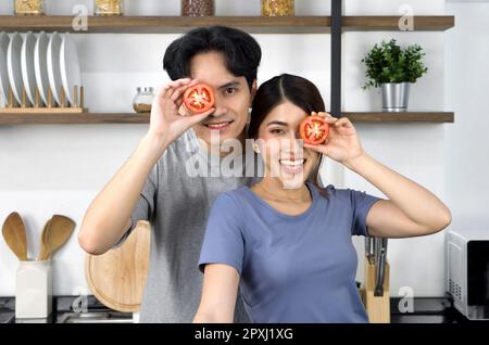 Giovane coppia in vestiolo casual, tenere il pomodoro a fette in posizione occhio. Gli amanti asiatici trascorrono il tempo di mattina insieme in cucina. Foto Stock