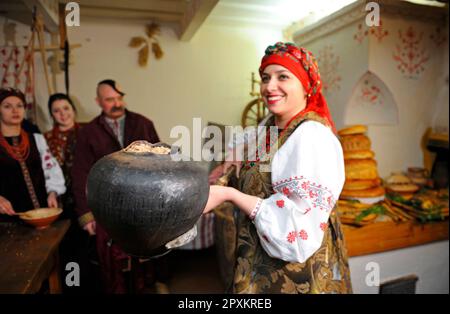 Donna in ucraino abito nativo utilizzando dispositivo 'rogach' per cucinare piatti tradizionali kutia in un forno rustico. Ricostruzione delle tradizioni popolari ucraine. JA Foto Stock