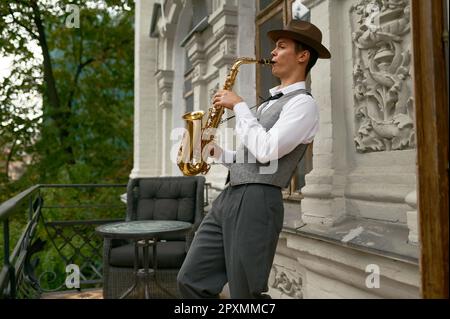 Giovane musicista maschile che pratica sassofono in balcone a casa. Sassofonista ispirato che suona musica Foto Stock