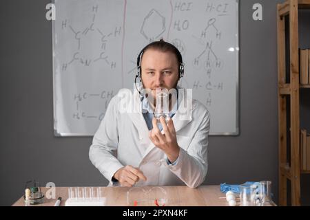 un insegnante di chimica al barbuto con una cuffia in bianco siede su una sedia sullo sfondo di un cartello con le formule, tenendo una fiasca di liquido Foto Stock