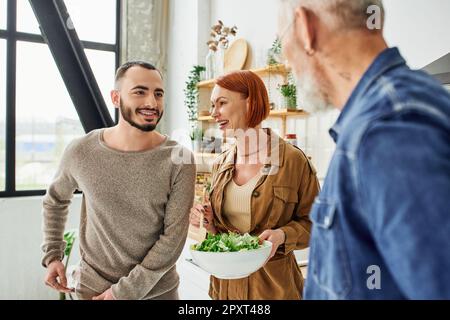 felice donna arrethead tenere ciotola con insalata fresca vicino al figlio e sfocato marito in cucina, immagine stock Foto Stock