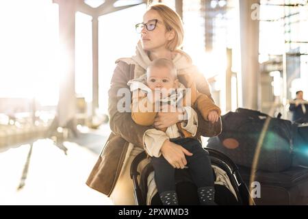 Motherat viaggia con il bambino, camminando, spingendo passeggino e carretto bagagli davanti alla stazione del terminal aeroportuale Foto Stock