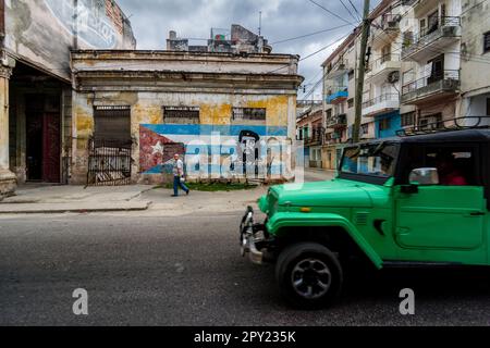 Una jeep verde che si muove attraverso una strada a l'Avana con una bandiera cubana e un uomo che cammina sullo sfondo principale fuoco, la tipica architettura dell'Avana Foto Stock