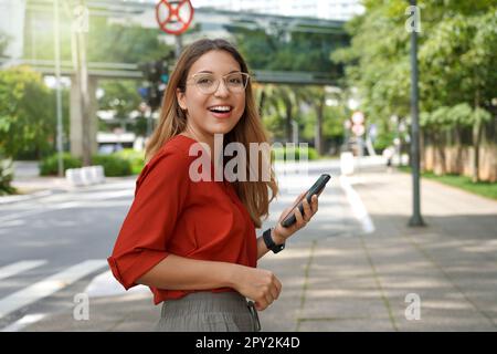 Donna brasiliana vivace e fiduciosa sorridente alla fotocamera che tiene lo smartphone nella moderna metropoli sostenibile Foto Stock