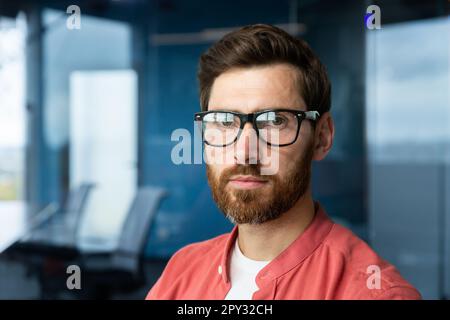 Primo piano. Ritratto di un giovane professionista, di uno sviluppatore in occhiali e di una camicia rossa che guarda con fiducia nella fotocamera. Foto Stock