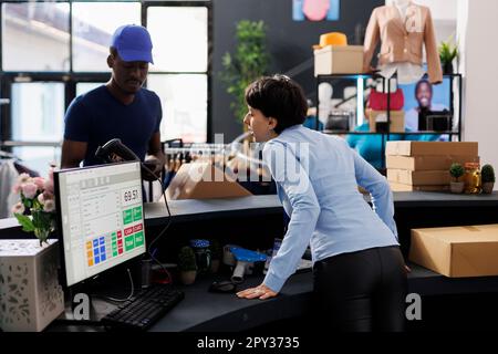 Lavoratore caucasico discutere con corriere, preparare i pacchetti per la spedizione in boutique moderna. Dipendente in piedi al banco, che controlla il report logistico sul tablet. Concetto di moda Foto Stock