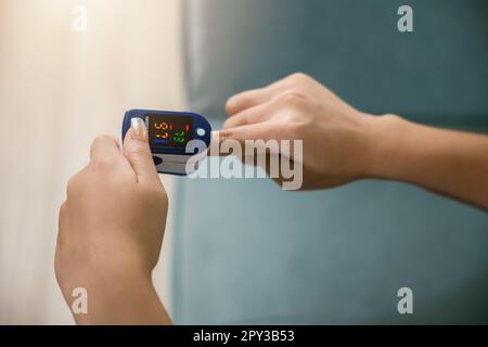 Giovane donna che usa il pulsossimetro per monitorare i livelli di ossigeno nel sangue e la frequenza del polso sul dito sotto il divano nel soggiorno a casa, Self healthcare con coron Foto Stock