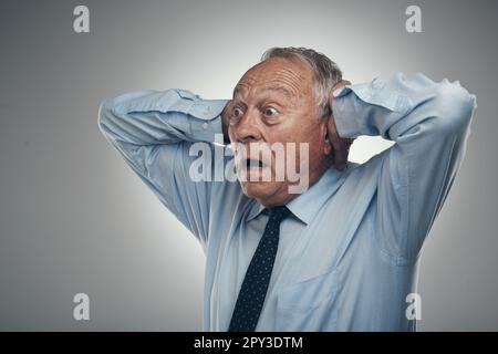 OH, è forte. un uomo d'affari anziano in piedi contro uno sfondo grigio studio con la testa nelle sue mani e guardando scioccato. Foto Stock