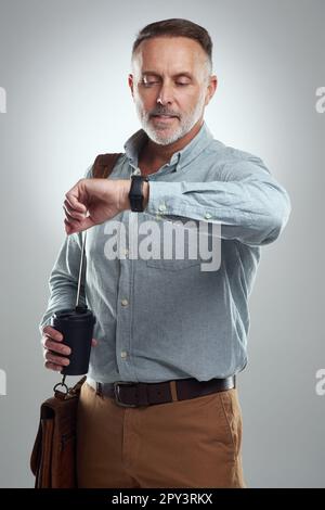 Im puntualmente, come previsto. Studio shot di un uomo maturo che porta una borsa e una tazza di caffè mentre il tempo sul suo orologio da polso su uno sfondo grigio Foto Stock