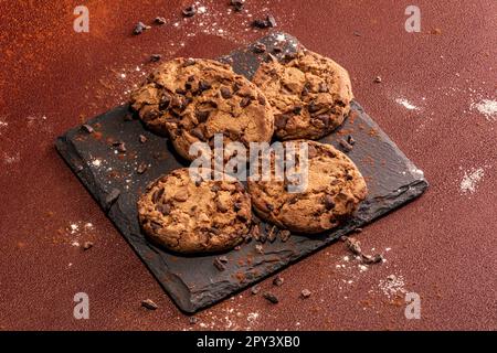 Cinque biscotti con gocce di cioccolato su una lastra di ardesia su uno sfondo marrone dipinto a mano. Atmosfera rustica. Foto Stock