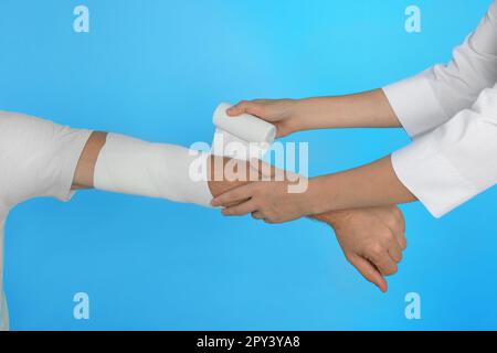 Medico che applica la bendatura medica sul braccio del paziente su sfondo azzurro, primo piano Foto Stock