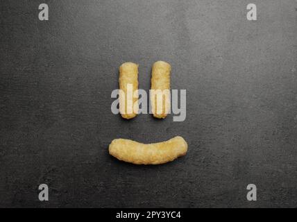 Faccia divertente fatta di bastoni di mais su tavolo grigio scuro, piatto Foto Stock
